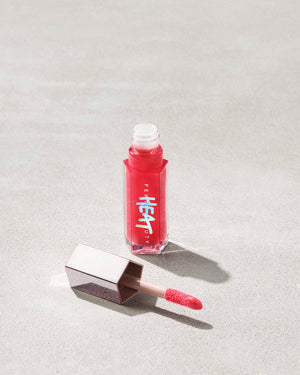 An opened tube of Gloss Bomb Heat lip gloss and plumpler in the shade Hot Cherry on a grey background.