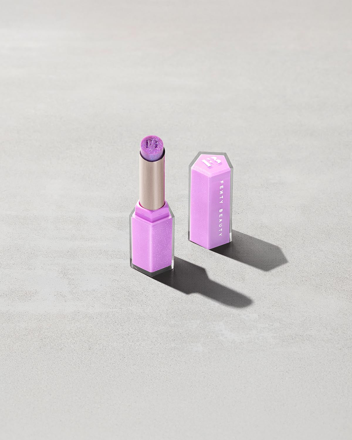 Open product shot of Gloss Bomb Stix Shimmer in shade Ice Amethyst on a concrete background.