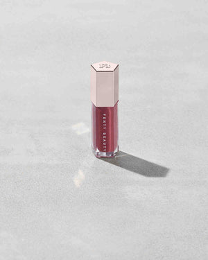 A closed tube of Gloss Bomb Universal Lip Luminizer in the shade RiRi on a grey background.