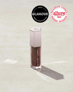A closed tube of Gloss Bomb Universal Lip Luminizer in the shade Hot Chocolit on a grey background.