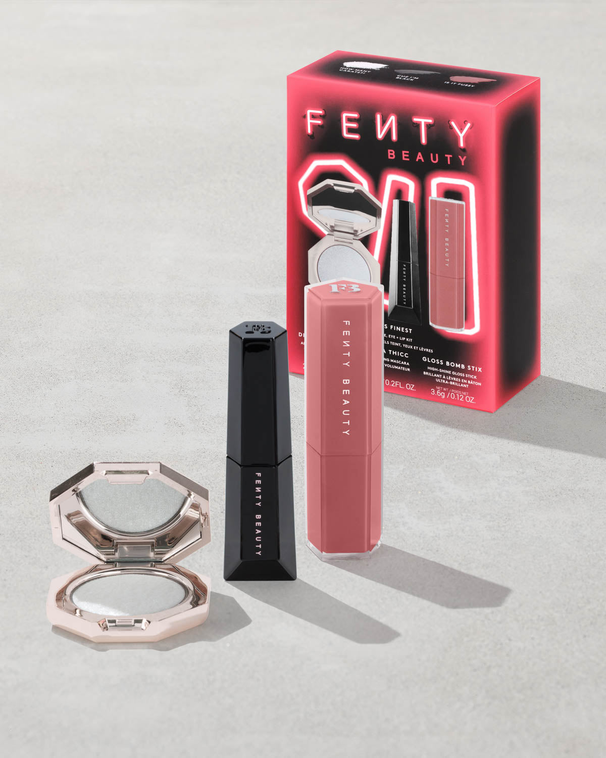 Fenty Beauty makeup items including gloss bomb stix in Is it Fu$$y, diamond bomb with a compact mirror, and a mini mascara on a light gray concrete background