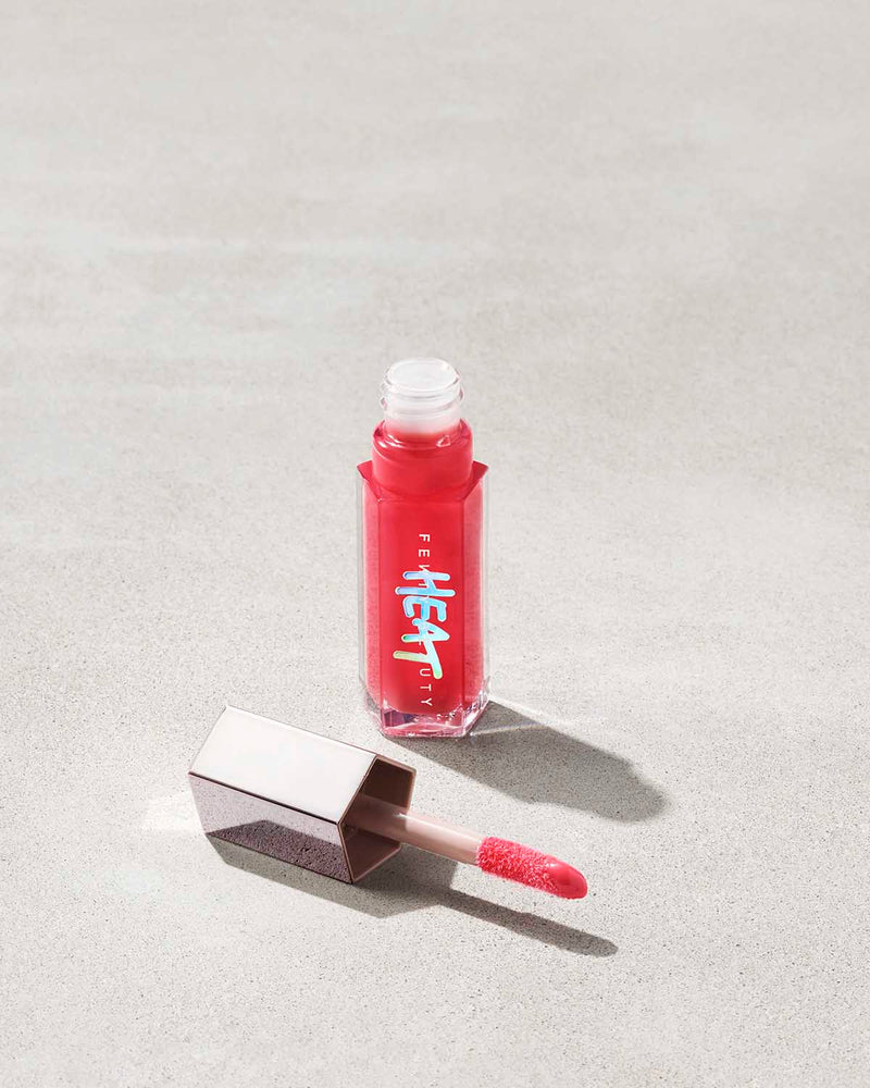 An opened tube of Gloss Bomb Heat lip gloss and plumpler in the shade Hot Cherry on a grey background.