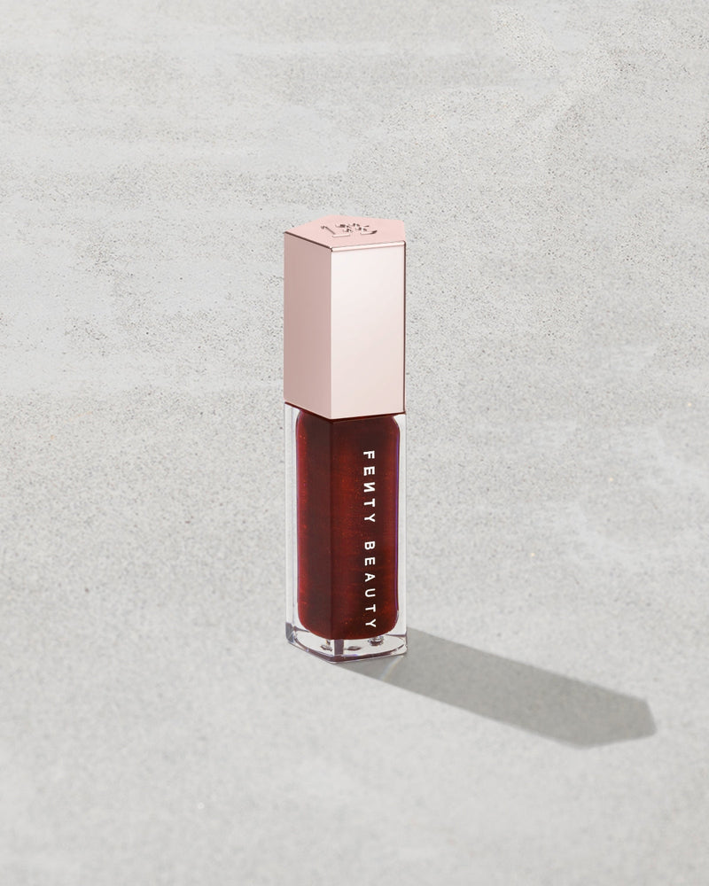 Fenty Beauty liquid lipstick in a deep berry red shade, housed in a clear bottle with a metallic rose gold cap, set against a textured light gray background.